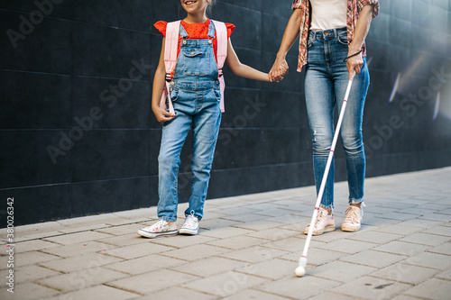 Young blind mother walking with her little daughter on city street. They wearing face protective masks. Back to school and new coronavirus lifestyle concept. photo