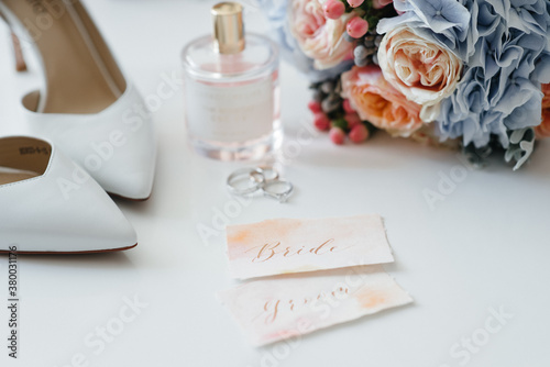 Wedding accessory bride. Stylish beige shoes, earrings, gold rings, flowers, garter, perfumes on table standing on wooden background. Letters from the bride and groom. flat lay. top view.