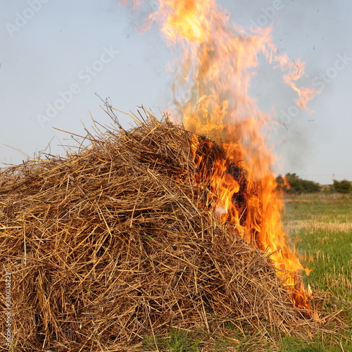 Fotografie, Tablou Fire in the grass pile, burning straw in haystack on field and blue sky backgrou