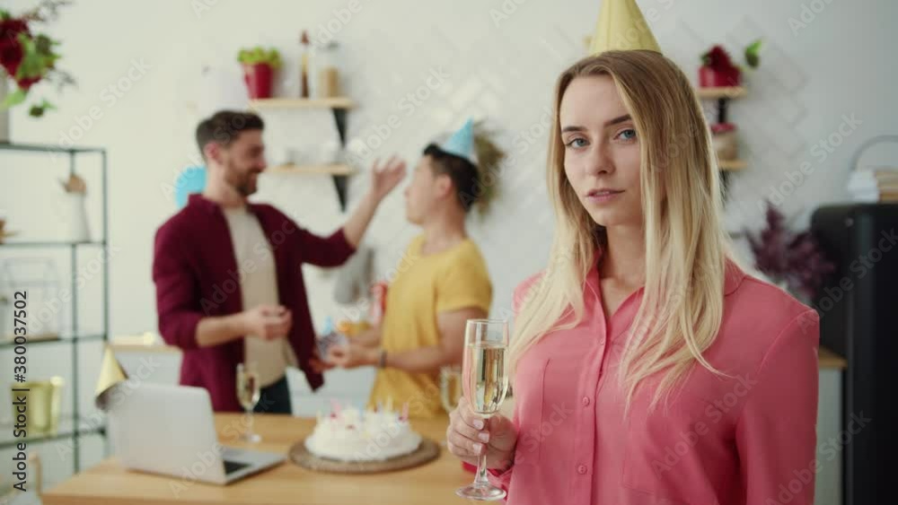 Blonde caucasian beautiful girl with glass of champagne smiling at camera. In background multi-race gay couple wearing birthday hats preparing holiday party. Blonde caucasian beautiful girl with glass of champagne smiling at camera. In background multi-race gay couple wearing birthday hats preparing holiday party.