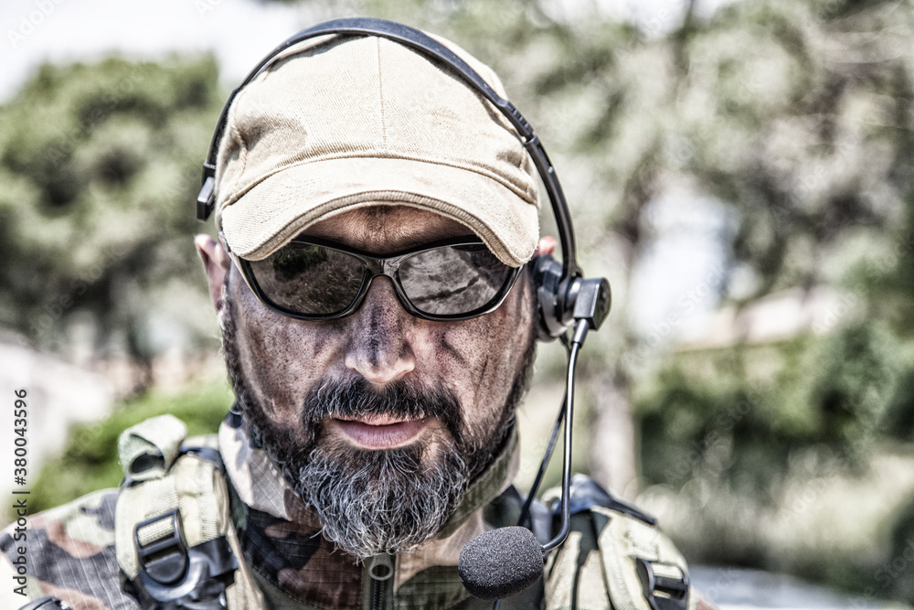 Shoulder portrait of army soldier, special forces fighter, modern warfare combatant with dirty, unshaven face, wearing sunglasses, combat helmet and talking in tactical radio headset during mission Shoulder portrait of army soldier, special forces fighter, modern warfare combatant with dirty, unshaven face, wearing sunglasses, combat helmet and talking in tactical radio headset during mission