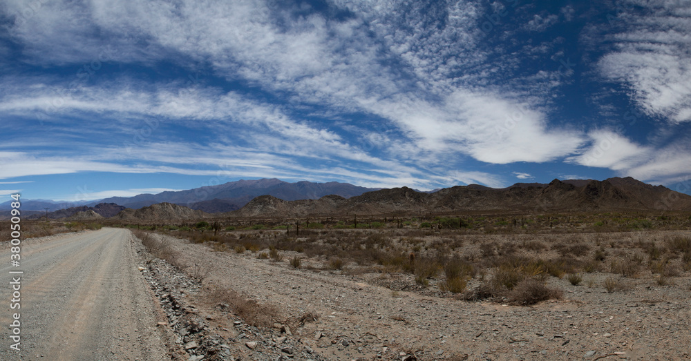 Traveling along the dirt road. Panorama view of the empty route across the arid desert and mountains under a beautiful blue sky with clouds. 