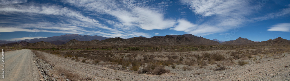 Traveling along the dirt road. Panorama view of the empty route across the arid desert and mountains under a beautiful blue sky with clouds.