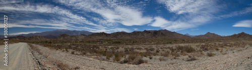 Traveling along the dirt road. Panorama view of the empty route across the arid desert and mountains under a beautiful blue sky with clouds. © Gonzalo