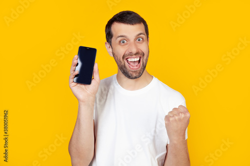 A young man with a beard in a white T-shirt shows a smartphone screen, shouts with pride and celebrates victory and success, is very excited, rejoices in emotions. Stands on isolated yellow background