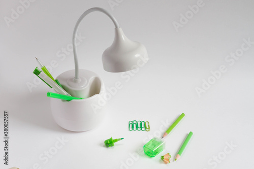 Green stationery, pencil, pen, felt-tip pen, sharpener, paper clips are on the white table by the table lamp. Beautiful lamp with a compartment for storing office supplies.