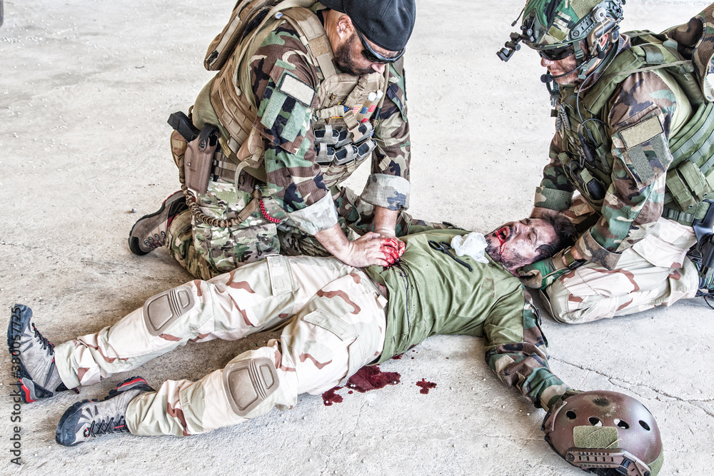 Soldiers trying to stop bleeding at wounded comrade who lying on floor, suffering and screaming in paine. Commando fighter pressing with hands on bloody wound at friends stomach, giving emergency care Soldiers trying to stop bleeding at wounded comrade who lying on floor, suffering and screaming in paine. Commando fighter pressing with hands on bloody wound at friends stomach, giving emergency care