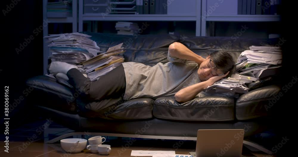 Tired senior asian business man is sleeping on sofa covered with stack of paperwork and have laptop, cup of coffee lay on the floor. Alone male fall asleep on couch while working hard at late night. Tired senior asian business man is sleeping on sofa covered with stack of paperwork and have laptop, cup of coffee lay on the floor. Alone male fall asleep on couch while working hard at late night.