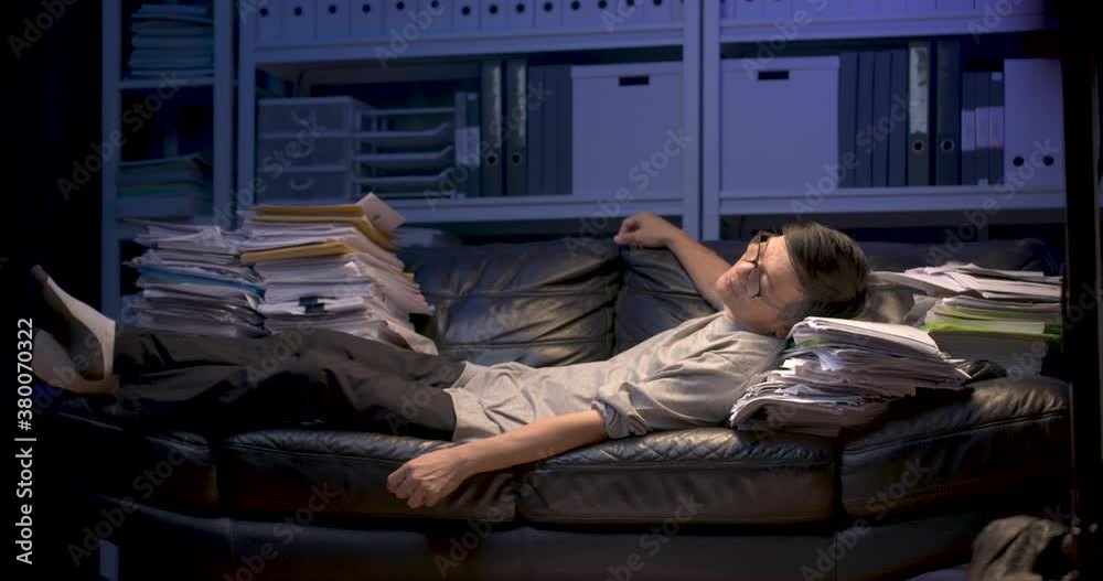 Tired senior asian man is sleeping on sofa covered with stack of paperwork while working hard at late night. Stressed alone male having restless sleep, feeling uncomfortable in couch. Dolly shot