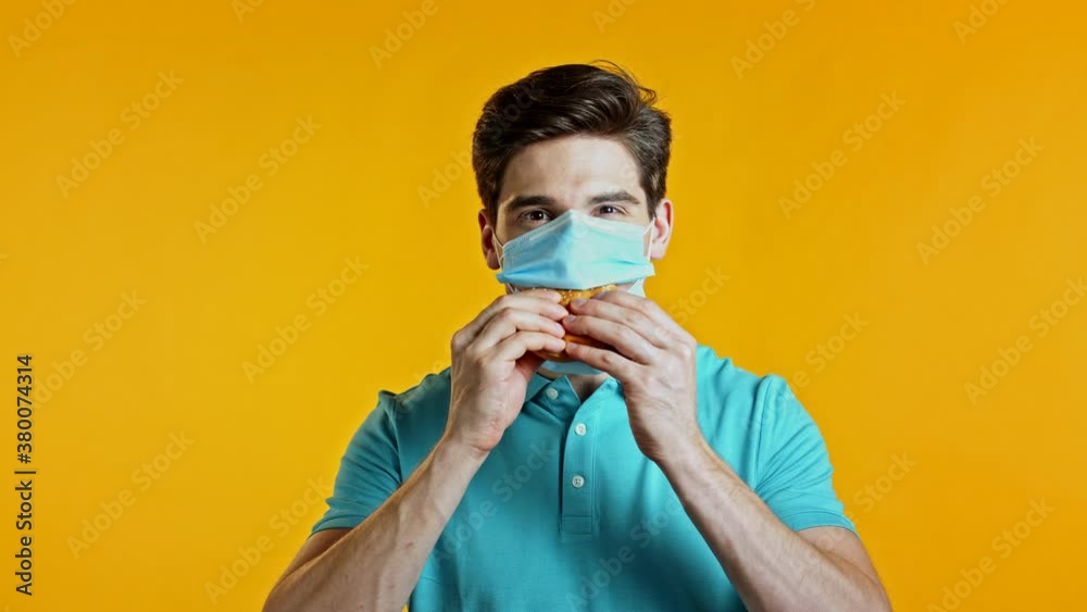 Young man in protective medical mask eating burger on yellow studio background. Guy makes fun, shows how to live during an epidemic. Young man in protective medical mask eating burger on yellow studio background. Guy makes fun, shows how to live during an epidemic.