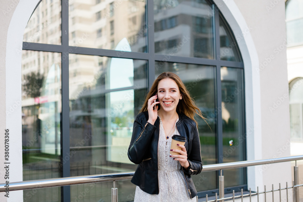 Beautiful woman is speaking on the phone and smiling, with a cup of coffee in the hand, against the glass facade in the city Beautiful woman is speaking on the phone and smiling, with a cup of coffee in the hand, against the glass facade in the city