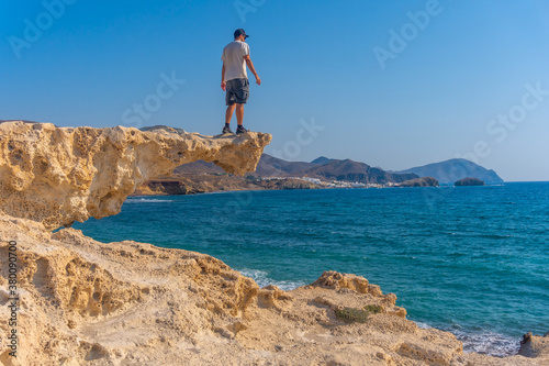 A young man perched on the rocks looking at Playa los Escullos in the natural park of Cabo de Gata, Nijar, Andalucia. Spain, Mediterranean Sea