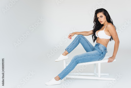 Fotografering brunette woman in denim jeans and bra sitting on stool on grey