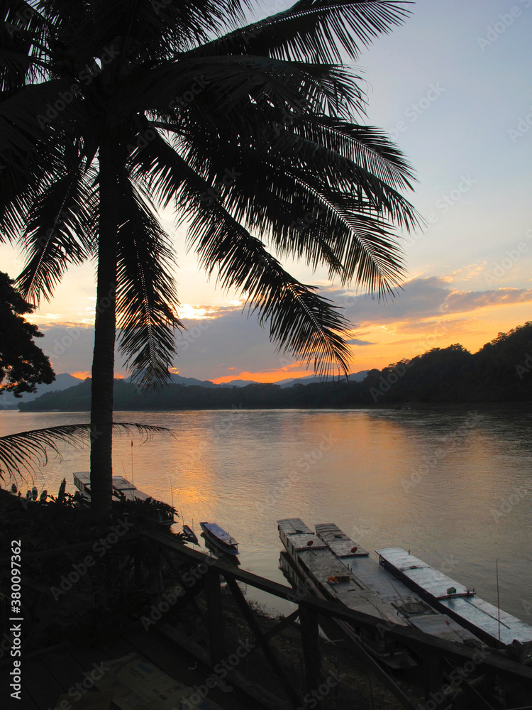 Fototapeta premium Scenic sunset view over the Mekong River in Laos, with palm trees and traditional wooden rafts and boats
