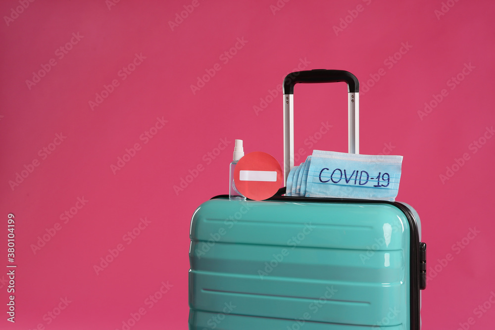 Antiseptic spray, stop sign and protective masks with inscription COVID-19 on suitcase against pink background, space for text. Travel restriction during coronavirus pandemic Antiseptic spray, stop sign and protective masks with inscription COVID-19 on suitcase against pink background, space for text. Travel restriction during coronavirus pandemic