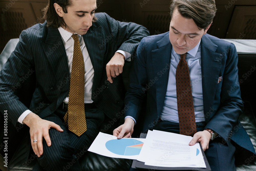 Two Businessmen Sat On Couch Reviewing Documents Two Businessmen Sat On Couch Reviewing Documents