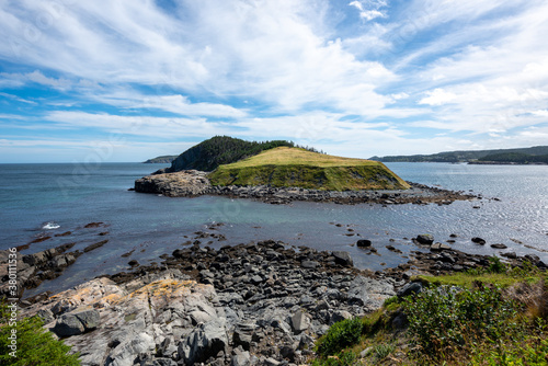 A small island surrounded by the blue ocean. The island has a grassy knoll and a small hill covered in trees. The sky is blue with dramatic clouds. There's a steep rocky cliff with beach boulders.