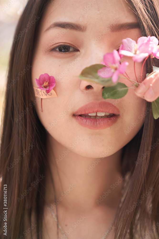 Portrait of a beautiful Asian woman in a white dress, she stands near a tree with white flowers, on her cheeks pink flowers with petals glued to a band-aid, she looks directly at the camera Portrait of a beautiful Asian woman in a white dress, she stands near a tree with white flowers, on her cheeks pink flowers with petals glued to a band-aid, she looks directly at the camera