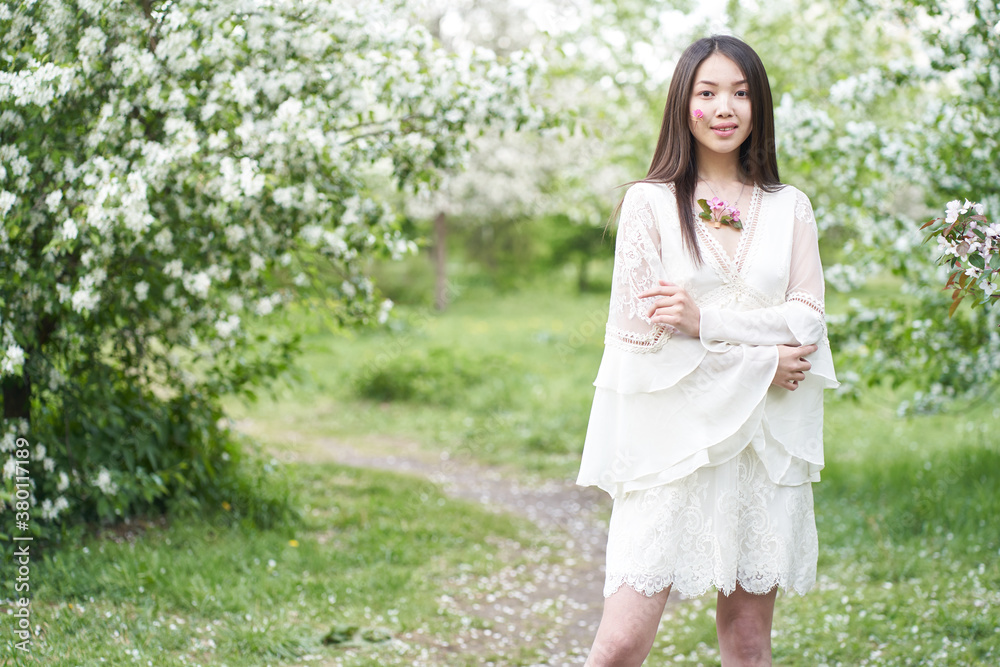 A beautiful Asian woman is standing near a tree with white flowers, pink petals on her cheeks, looking directly at the camera A beautiful Asian woman is standing near a tree with white flowers, pink petals on her cheeks, looking directly at the camera