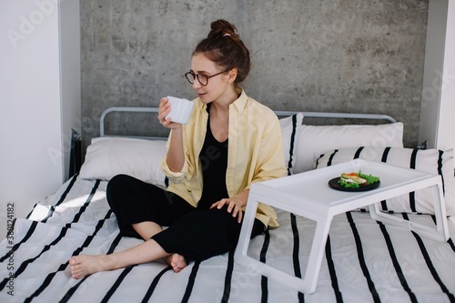 Young female wearing glasses taking breakfast in the bed and drinking tea or coffee photo