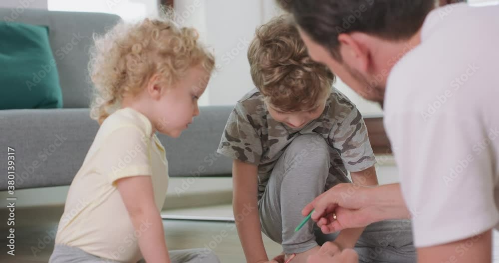 Close up of a little boy and a little girl, and their father are drawing with pencils on the white paper on the floor. Father explains something to the boy, who is looking atttentively at the father Close up of a little boy and a little girl, and their father are drawing with pencils on the white paper on the floor. Father explains something to the boy, who is looking atttentively at the father