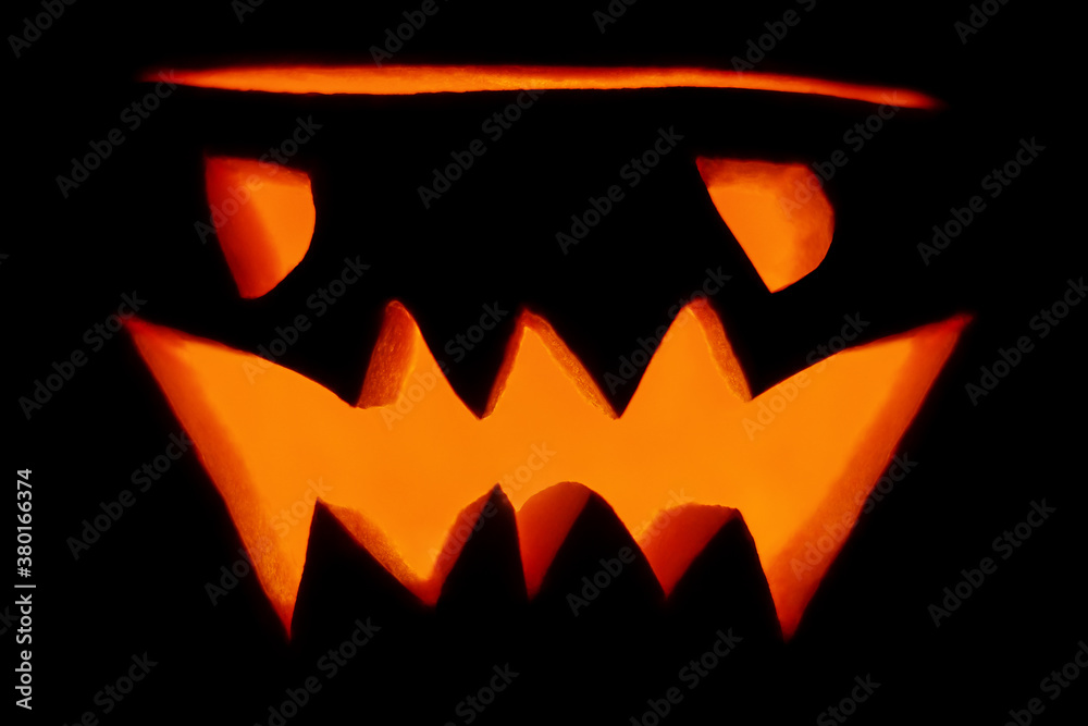 Fototapeta premium Evil orange glowing lantern-Jack carved from a pumpkin for the holiday Halloween close-up in the dark isolated on a black background. Ugly pumpkin face with a burning candle inside