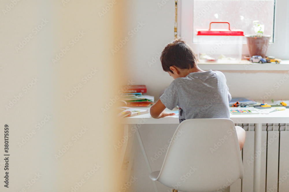 dark-haired boy draws a picture with pencils while sitting at a white table dark-haired boy draws a picture with pencils while sitting at a white table