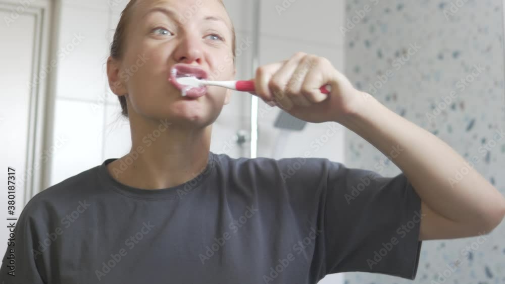 A young woman actively brushes her teeth in the morning, reflected in the mirror in the bathroom.