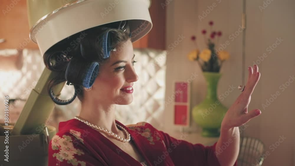 Medium shot of a woman sitting in her house with a mid-century modern retro style hood dryer over her head, looking at her nails. Medium shot of a woman sitting in her house with a mid-century modern retro style hood dryer over her head, looking at her nails.