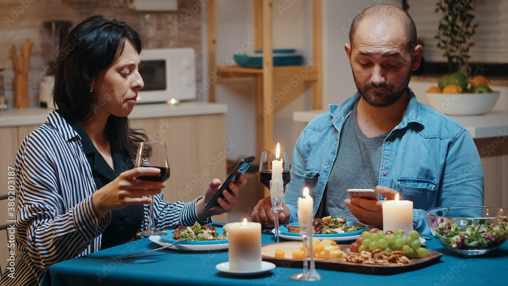Focused married couple reading news of phones during romantic dinner. Adults sitting at the table, browsing online, surfing, using smartphones, celebrating their anniversary in the dining room. Focused married couple reading news of phones during romantic dinner. Adults sitting at the table, browsing online, surfing, using smartphones, celebrating their anniversary in the dining room.