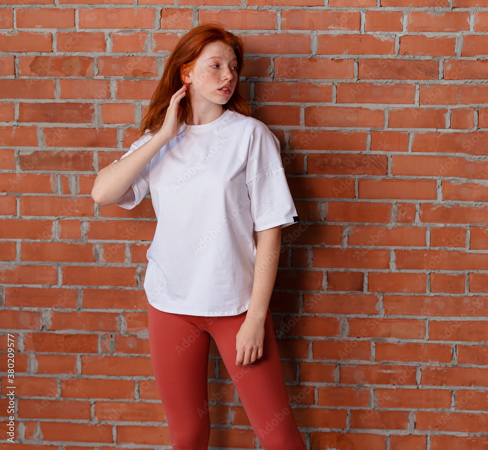 Fashion style studio portrait of beautiful red-haired young girl in leggings pants and white t-shirt. Model standing and posing beside brick wall Fashion style studio portrait of beautiful red-haired young girl in leggings pants and white t-shirt. Model standing and posing beside brick wall