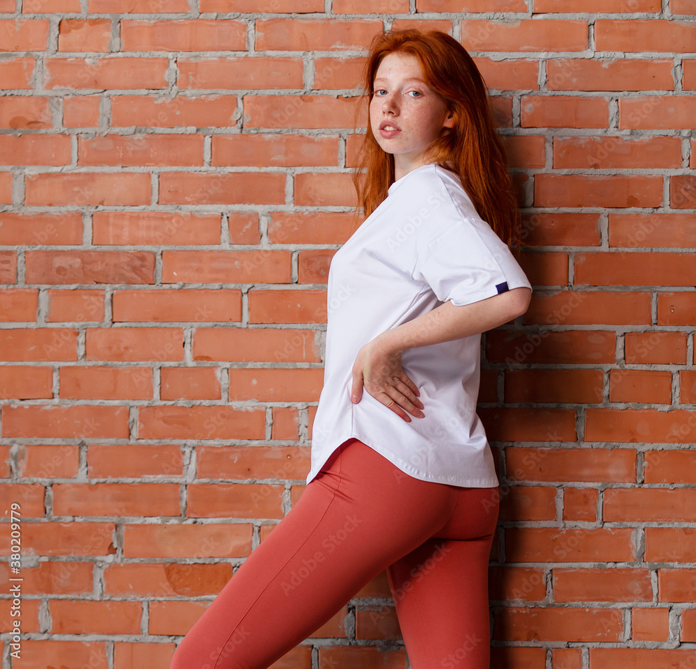 Fashion style studio portrait of beautiful red-haired young girl in leggings pants and white t-shirt. Model standing and posing beside brick wall Fashion style studio portrait of beautiful red-haired young girl in leggings pants and white t-shirt. Model standing and posing beside brick wall