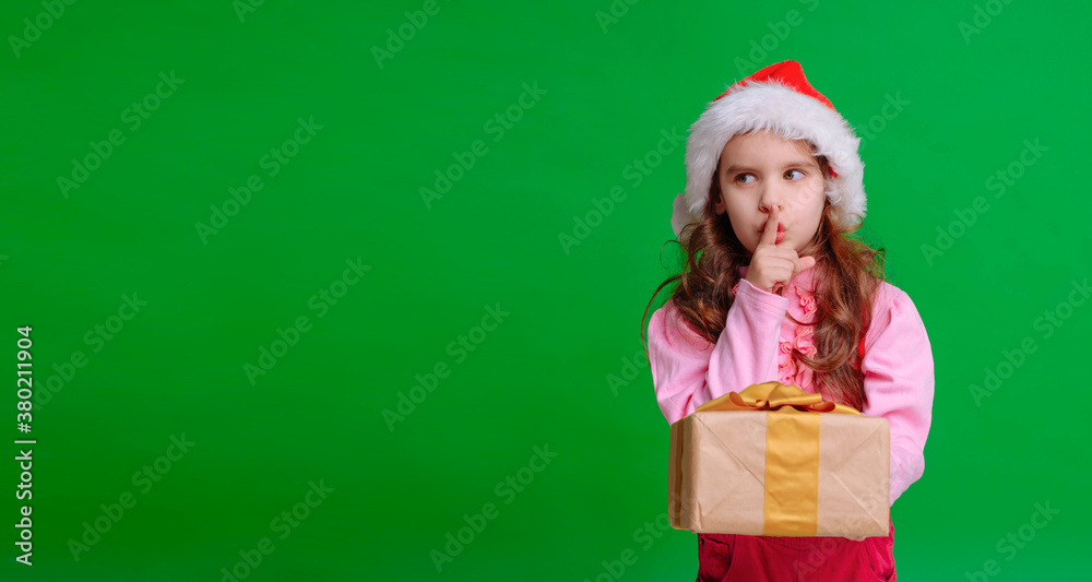 Cute girl in a Santa hat holds a Christmas gift isolated on a green background. Cute girl in a Santa hat holds a Christmas gift isolated on a green background.