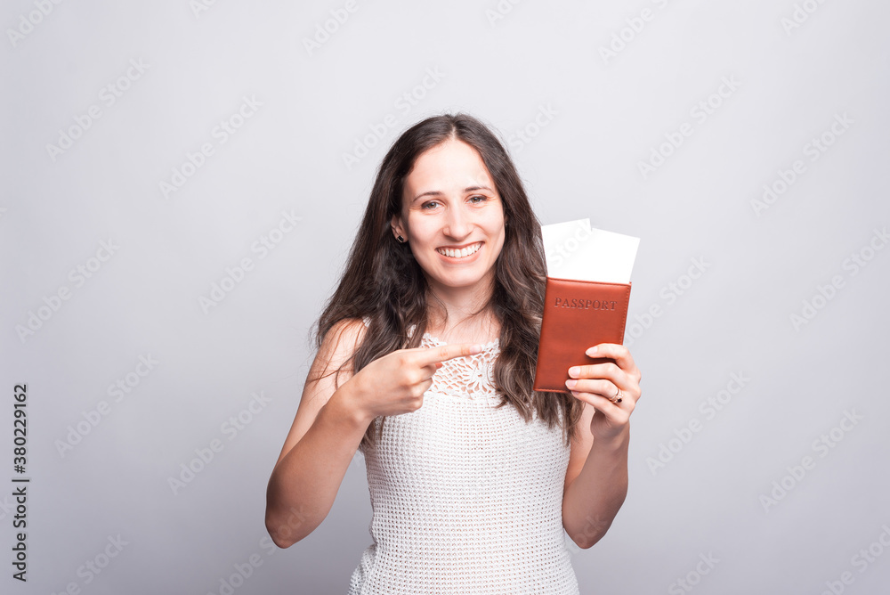 Happy young woman is holding a passport and with some tickets in it is smiling and looking at the camera near a gray wall Happy young woman is holding a passport and with some tickets in it is smiling and looking at the camera near a gray wall