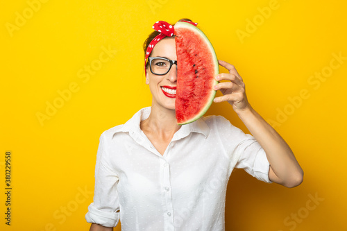 Young woman in a red headband and glasses covers half of her face with watermelon on a yellow background