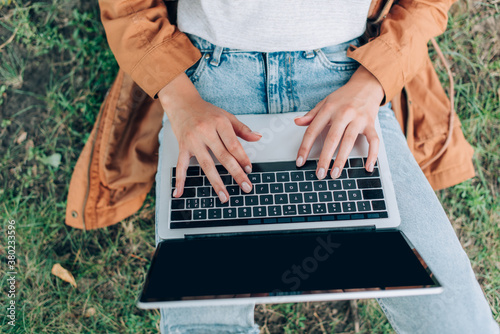 Top view of teleworker in raincoat using laptop with blank screen on grassy lawn