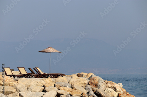 Fototapeta Naklejka Na Ścianę i Meble -  Empty deck chairs and sun umbrella on a stone beach on misty mountains background. Beach vacation on a sea	