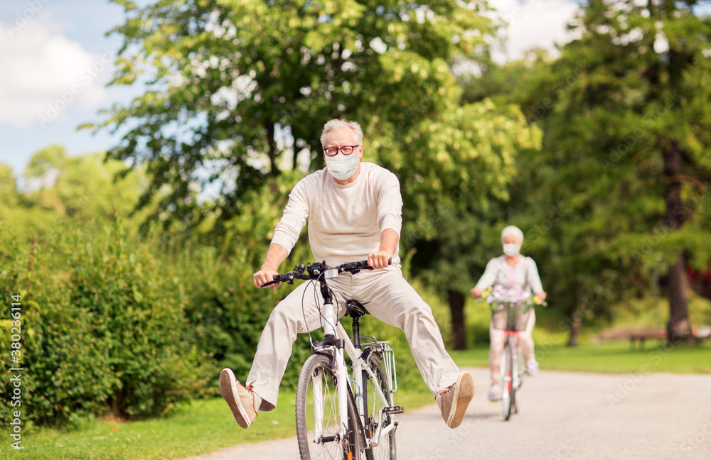 old age, health and pandemic concept - happy senior couple wearing face protective medical mask for protection from virus disease riding bicycles at summer park