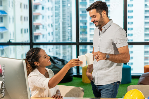 Two business partnership Caucasian man with Hispanic woman taking a break and enjoy drinking coffee together at office. Happy young creative people office worker colleague talking about work project.