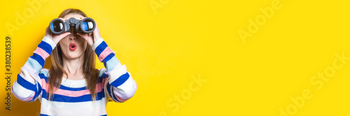 Young woman with a surprised face looks through binoculars on a yellow background. Banner