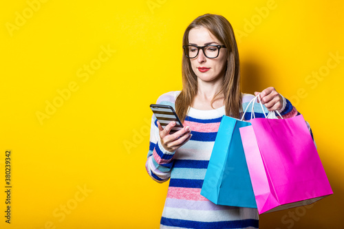 Young woman looks into the phone and holds shopping bags with purchases on a yellow background. Banner