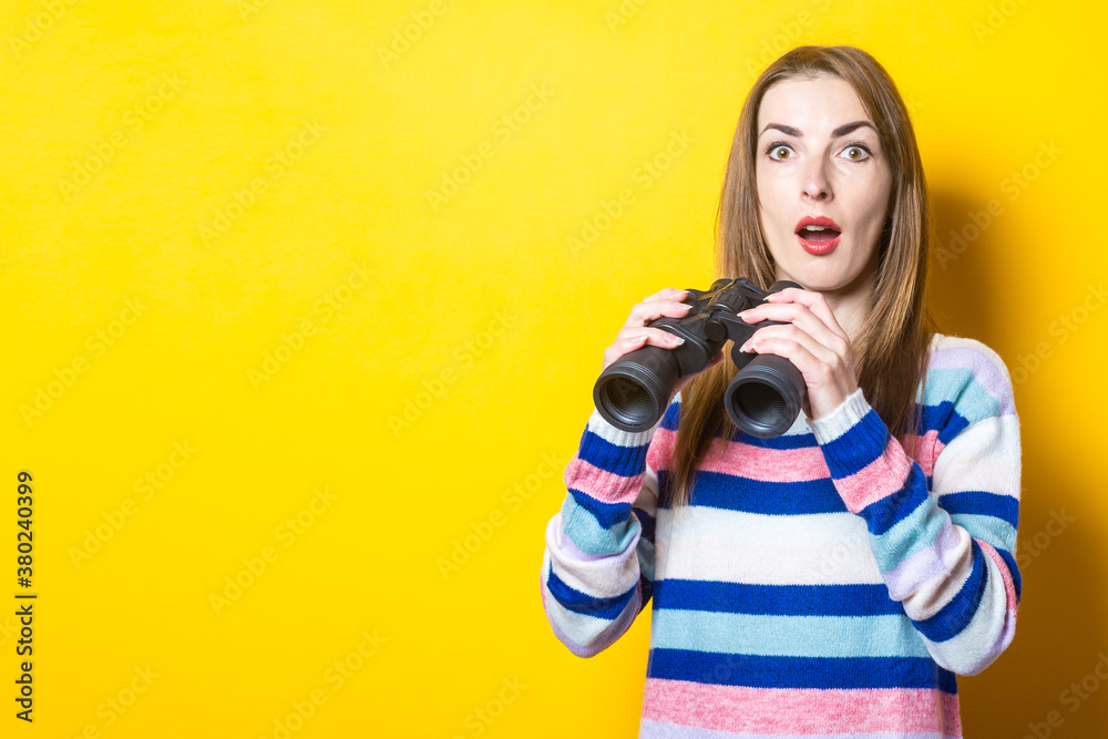Young woman with a surprised face holds binoculars on a yellow background. Banner Young woman with a surprised face holds binoculars on a yellow background. Banner