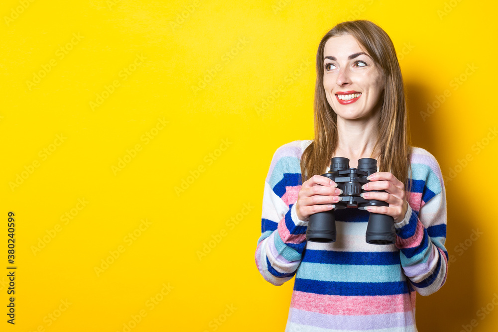 Young woman with a smile holds binoculars in her hands on a yellow background. Banner Young woman with a smile holds binoculars in her hands on a yellow background. Banner