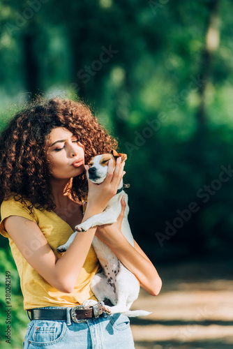 curly woman in summer outfit making duck face while holding jack russell terrier dog in park