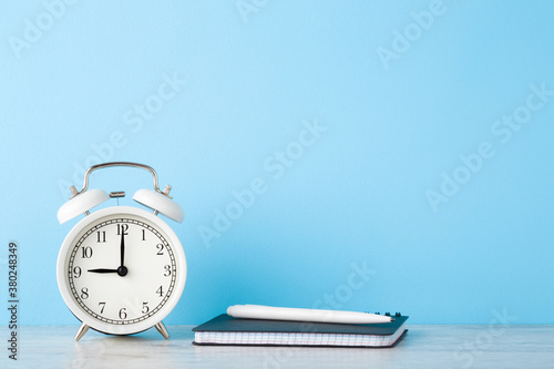 White alarm clock, planner with pen on wooden table at light blue wall background. Planning time concept. Closeup. Empty place for text. Front view.