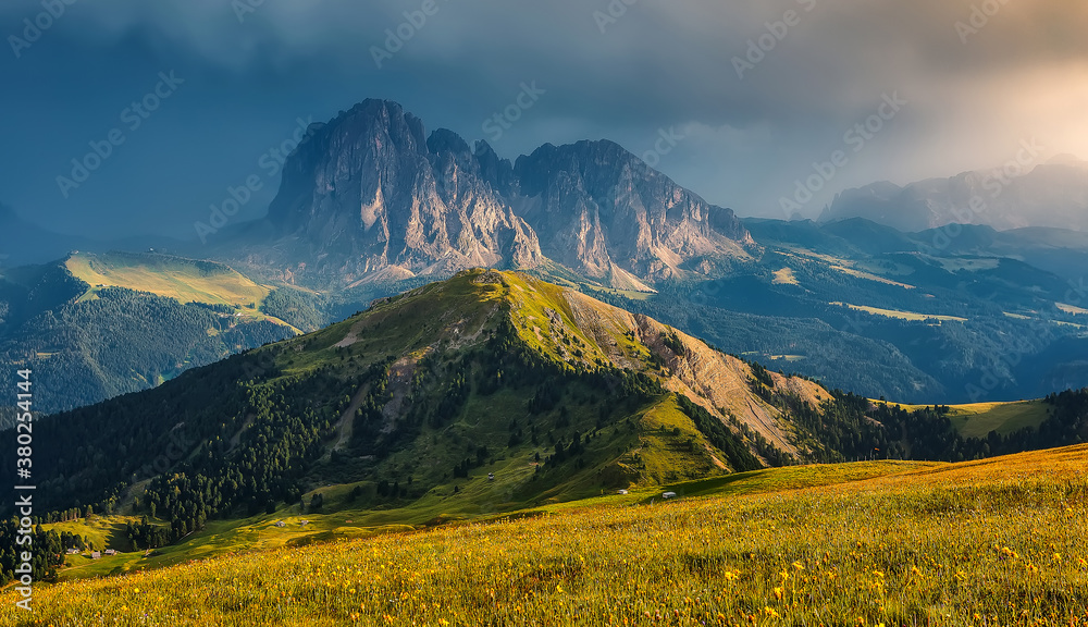 Scenic panoramic view of mountain hills landscape with blooming meadows and majestic alpine mountain peaks in the background on a colorful sunset with overcast sky in summer. amazing nature scenery