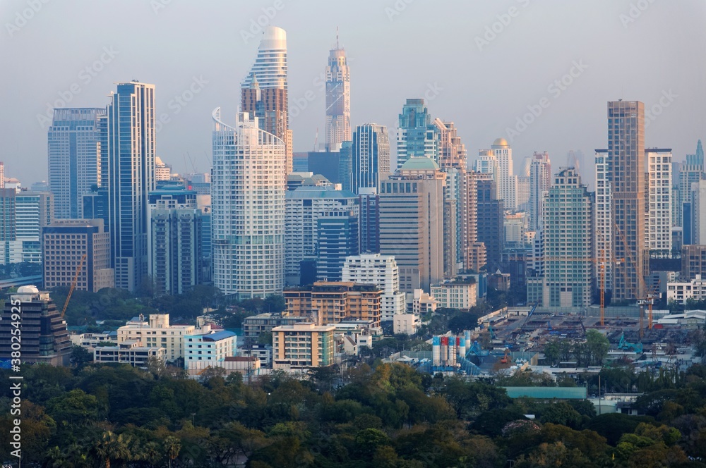 Fototapeta premium Urban skyline of Bangkok, the vibrant capital city of Thailand, with modern high rise skyscrapers in background and treetops of forests in beautiful Lumphini Park in foreground in early morning light