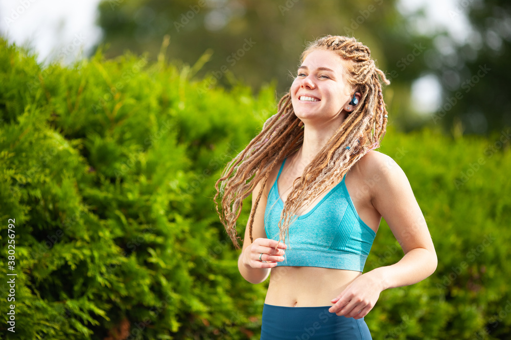 Athletic girl with dreadlocks running outdoors by the green fence. Sport and healthy lifestyle concept. In headphones.. Athletic girl with dreadlocks running outdoors by the green fence. Sport and healthy lifestyle concept. In headphones..