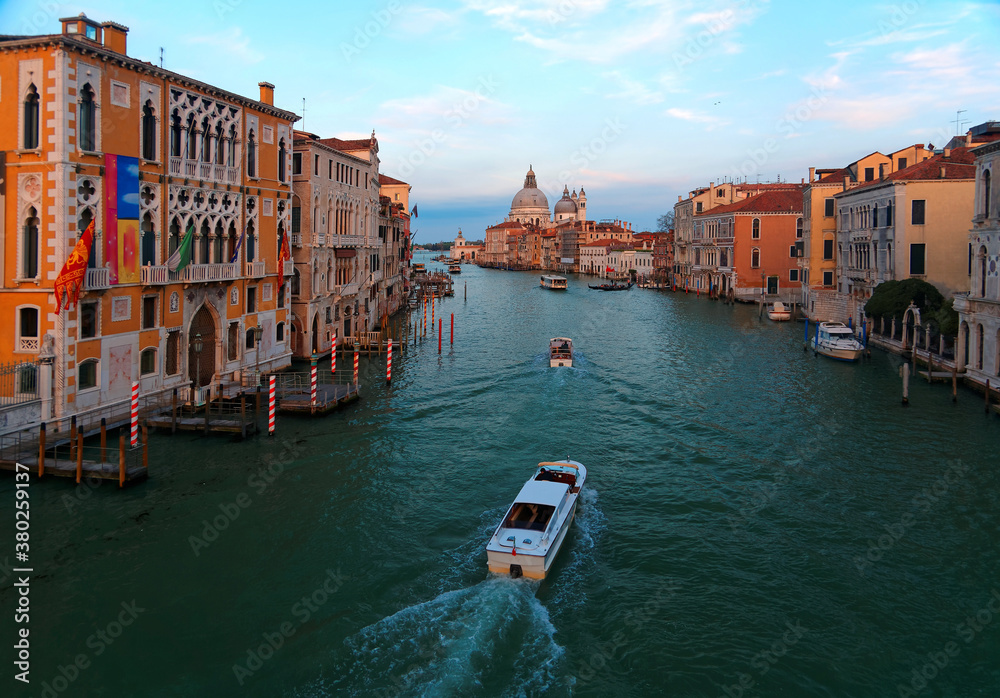 Fototapeta premium Romantic scenery of Venice at sunset, with boats, ferries & gondolas cruising on Grand Canal & the majestic landmark building Basilica di Santa Maria della Salute bathed in warm sunlight in background