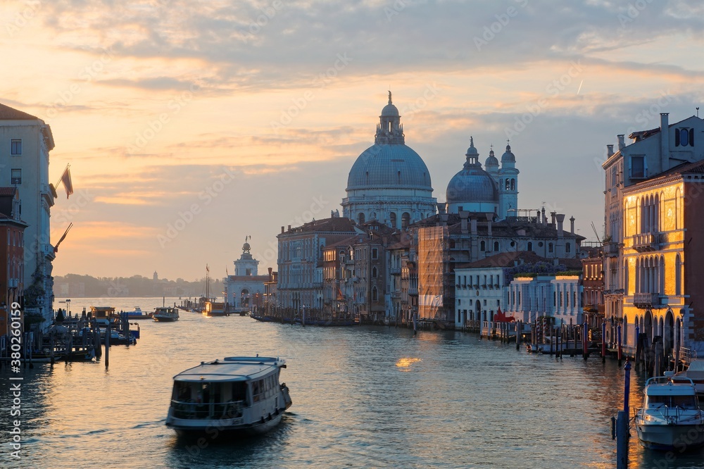 Morning scenery of the Grand Canal bathed in golden twilight, with boats & ferries cruising on waterway & landmark cathedral Basilica Santa Maria della Salute in background, in romantic Venice, Italy